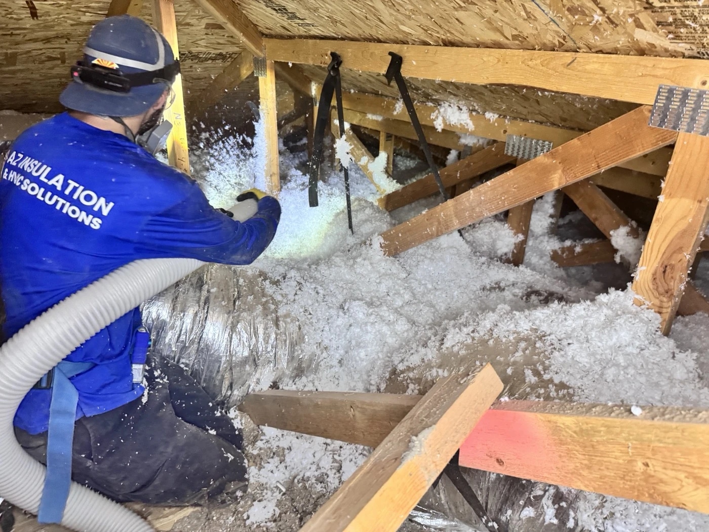 An AZ Insulation technician blowing loose-fill fiberglass around trusses and ductwork in an Arizona attic.